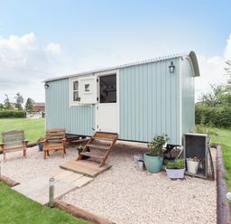 A small light blue wooden cabin with white door and window on a gravel area with two wooden chairs and potted plants at Honeybee Lodge in Stixwould near Woodhall Spa