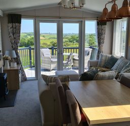 A living room with sofa and cushions a wooden dining table and chairs and glass doors leading to a balcony with outdoor seating at Vendee Lodge Thorness Bay Holiday Park near Cowes