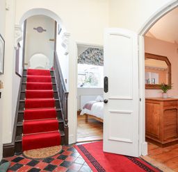 An entryway with a staircase covered by a red carpet a bedroom visible through a doorway and a wooden cabinet with a mirror in a living room at Ty Biwmares in Beaumaris