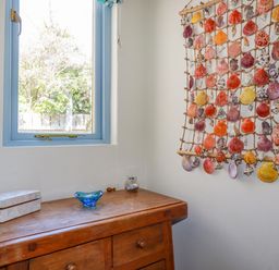 A wooden dresser under a window with a decorative box and glass items next to a wall hanging of seashells at Periwinkle Atlantic Highway near Bude