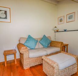 A seating area with a two-seater wicker sofa with cushions and matching footstool and wooden side tables at Periwinkle Atlantic Highway near Bude
