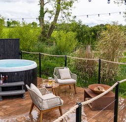 An outdoor patio with a hot tub two chairs a small table with drinks and snacks and a fire pit surrounded by greenery at Rusty Boathouse in Llanarth