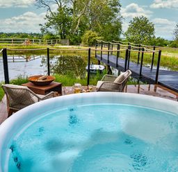 A hot tub with two chairs and a fire pit overlooking a pond with a wooden bridge at Rusty Boathouse in Llanarth