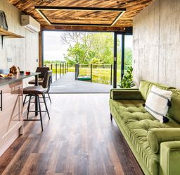 An open plan living space with a green sofa kitchen area with bar stools and a large sliding door opening to a deck overlooking water at Rusty Boathouse in Llanarth