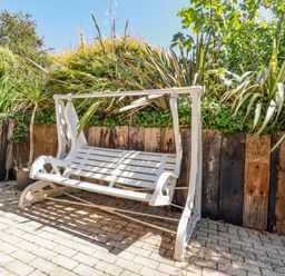 A white metal garden swing on a paved patio with wooden planter and leafy plants at Cameo Atlantic Highway near Bude