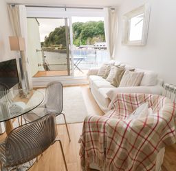 A living room with chairs a glass table a white sofa and a sliding glass door overlooking water at The Moorings in Porthmadog