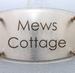 A metal and glass sign reading Mews Cottage mounted on a light blue textured wall at Mews Cottage in Laugharne