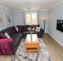 A living room with a gray sectional sofa, a wooden coffee table with flowers, a wall-mounted TV, and a floor lamp at Mews Cottage in Laugharne