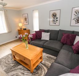 A living room with a dark grey sectional sofa with purple and white cushions a wooden coffee table with flowers a wooden cabinet with a lamp and framed leaf art on the walls at Mews Cottage in Laugharne