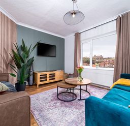 A living room with a blue sofa and brown armchair facing a TV on a wooden stand with a window showing a town view at The View in Whitby