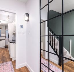 A hallway with a large grid mirror reflecting a staircase and a rug on wooden floor leading to a kitchen with white cabinets at The View in Whitby