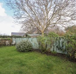 A garden with grass and bushes and a leafless tree and two white metal chairs next to a blue wooden fence
