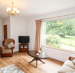 A living room with an armchair a sofa a coffee table a television and a large window showing green trees at Glebe Farm Cottage in Rathfriland County Down