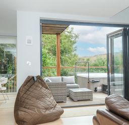 A seating area with a brown beanbag chair and leather sofa opening to an outdoor deck with wicker furniture and a hot tub at 9 Cedars in St Agnes
