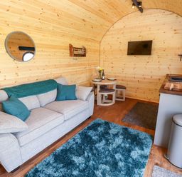 A wooden room with a sofa a round dining table a wall mirror a television and a kitchen area at The Happy Valley Pod in Chipping Norton