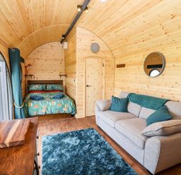 An interior of a wooden pod with a bed, sofa, rug, and curtains at The Happy Valley Pod in Chipping Norton