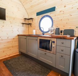 A kitchen area with grey cabinets a microwave a round window and wooden walls at The Happy Valley Pod in Chipping Norton