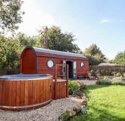 A small wooden cabin with round windows and a hot tub in a garden at The Happy Valley Pod in Chipping Norton
