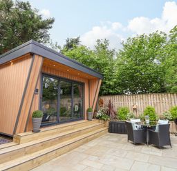 A wooden cabin with glass doors next to a patio with table and chairs and a barbecue at The Nest in Prestatyn