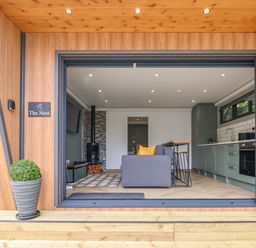 An open living room and kitchen with a gray sofa yellow cushions wood stove and green cabinets at The Nest in Prestatyn
