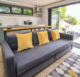 A kitchen area with green cabinets and white tiled backsplash with a gray sofa and yellow cushions in front at The Nest in Prestatyn