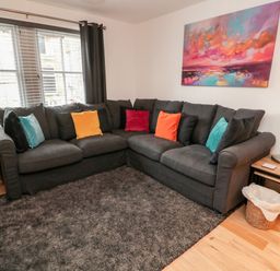 A living room with a dark gray sectional sofa with colorful pillows a side table with a lamp and plant and a painting on the wall at Lavenna in Alnwick