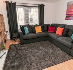 A living room with a dark gray corner sofa with colorful cushions a gray rug a wooden floor and a shelf with wicker baskets at Lavenna in Alnwick