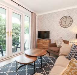 A living room with a sofa, armchair, nested coffee tables, a TV on a wooden cabinet, wall clock, lamp, and patio doors opening to outside at Bluebell Hill in Bowness-On-Windermere