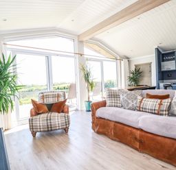 A living room with a plaid armchair and a leather sofa with cushions near large windows and plants at Atlantic Lodge Atlantic Highway near Bude