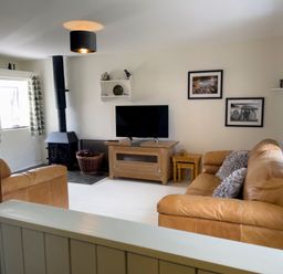 A living room with a tan sofa and armchair a television on a wooden stand a wood-burning stove and patterned curtains at The Shippen @ Canllefaes Penparc near Cardigan