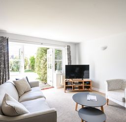 A living room with a sofa an armchair a TV on a stand and double doors leading to a garden at 12 The Gardens in Rousdon