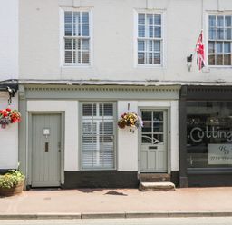 Shopfront with hanging flower baskets and two doors next to a hair salon with a union jack flag above the door at Cuckoo Cottage in Upton Upon Severn