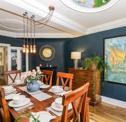 A dining room with a wooden table set for six with white dishes a blue vase with flowers wooden chairs a sideboard with a lamp and plant dark blue walls and a large framed painting at Cuckoo Cottage in Upton Upon Severn