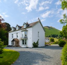 A white house with dormer windows and a gravel driveway surrounded by green trees and a grassy hill in the background at Swinside Lodge in Newlands Valley near Keswick