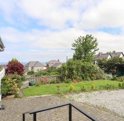 A garden with grass shrubs and trees next to a paved area and gravel driveway with houses in the background at Haddef Penmaenmawr in Penmaenmawr