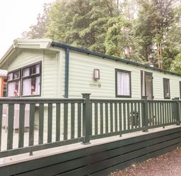 A light green static caravan with black framed windows and a dark green railing on a deck surrounded by trees at Herdwick Cabin in Troutbeck 68 White Cross Bay Holiday Park near Windermere
