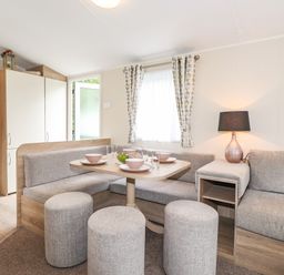 A dining area with a table set with bowls and glasses surrounded by upholstered bench seating and stools with a lamp on a side shelf by Herdwick Cabin in Troutbeck 68 White Cross Bay Holiday Park near Windermere