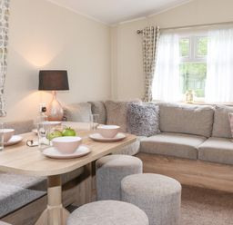 A dining area with a wooden table set with bowls and glasses surrounded by a grey upholstered corner sofa and round stools at Herdwick Cabin in Troutbeck 68 White Cross Bay Holiday Park near Windermere