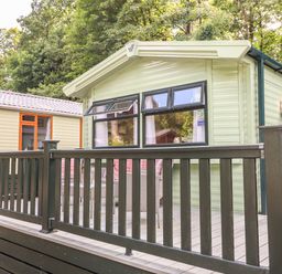 An outdoor deck with a black railing and patio furniture outside a green mobile home with windows and trees around at Herdwick Cabin in Troutbeck 68 White Cross Bay Holiday Park near Windermere