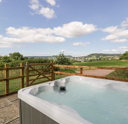 An outdoor hot tub on a stone patio next to a wooden fence with a countryside view at The Old Kennels in Harcombe near Sidford