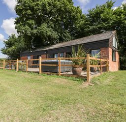 A single-story brick and wood building with a fenced patio and potted plants surrounded by grass and trees at The Old Kennels in Harcombe near Sidford