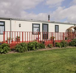 A single-story mobile home with windows door a red wooden deck and green plants in front with grass at The Cabin in Occumster near Lybster