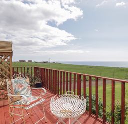 A wooden deck with two chairs and a table overlooking green fields and the sea at The Cabin in Occumster near Lybster