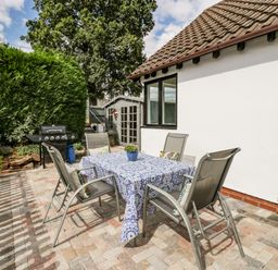 An outdoor patio area with a table covered by a blue patterned cloth and four chairs near a white house with a tiled roof and a barbecue grill at Phocle Ridge in Phocle Green near Ross-On-Wye