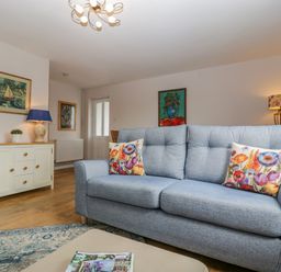 A living room with a blue sofa with floral cushions a sideboard with a lamp and sculpture and paintings on walls at Phocle Ridge Phocle Green near Ross-On-Wye