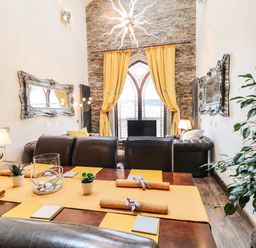 A dining area with a wooden table and brown leather chairs next to a living room with brown leather sofas and yellow curtains in front of a window at The Upper Room in Ballymena