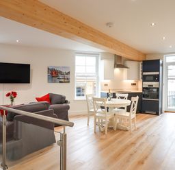 An open plan living room and kitchen with gray sofas red cushions a round dining table with four chairs and modern kitchen appliances at Ferryside Kingswear