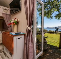 A small bedroom with bunk beds flowers on a cabinet and a wall-mounted tv opening onto a deck with a view of a grassy field and coastline at The Shepherd's Rest in Robin Hood's Bay
