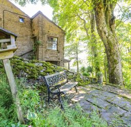 A stone house with a mossy stone wall a wooden birdhouse and a metal bench under large trees at Keepers in Nenthead near Alston