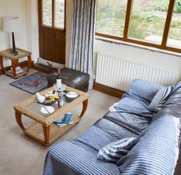 A living room with a leather sofa covered with blue striped throws a glass top coffee table with snacks a lamp on a wicker side table and a large window at Bothy in Nenthall near Alston